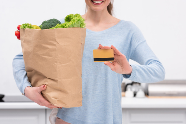 Schnappschuss einer lächelnden Frau mit Papiertüte und Kreditkarte in der Küche - Foto, Bild