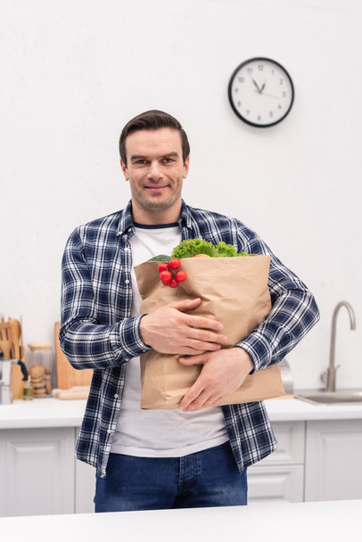 улыбающийся взрослый мужчина несет сумку из продуктового магазина на кухне и смотрит в камеру
 - Фото, изображение