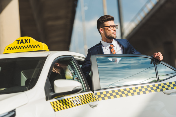 hansome young man in suit and eyeglasses looking away while sitting in taxi cab - Foto, Bild