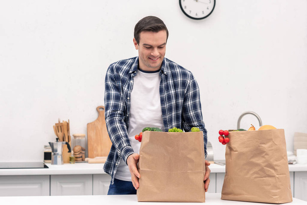 happy adult man with grocery store paper bags at kitchen - Foto, afbeelding