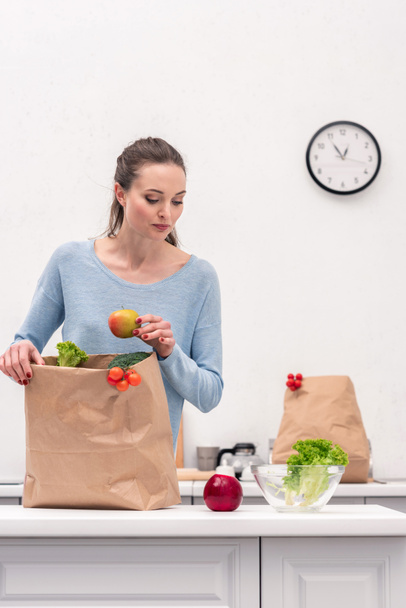 adult woman taking fruits and vegetables out of paper bag at kitchen - Foto, imagen