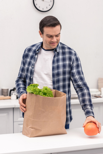 happy adult man unpacking grocery store paper bag at kitchen - Photo, Image