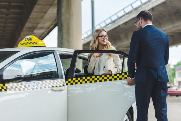 handsome stylish man opening car door to smiling blonde woman sitting in taxi - Zdjęcie, obraz