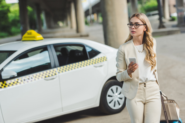 young woman in eyeglasses holding smartphone and looking away while standing near taxi cab on street - Photo, Image