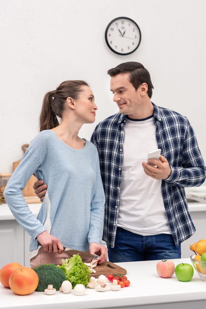 happy adult man with smartphone embracing his wife while she preparing dinner - Фото, изображение