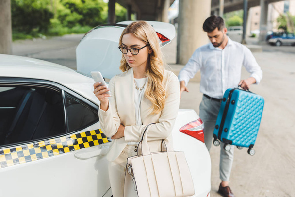 beautiful blonde woman in eyeglasses using smartphone while man putting suitcase in trunk of taxi car   - Fotó, kép