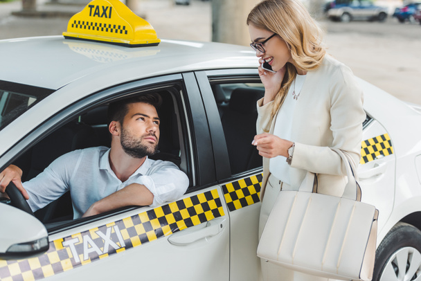 smiling blonde woman talking by smartphone and looking at male driver sitting in taxi - Photo, Image