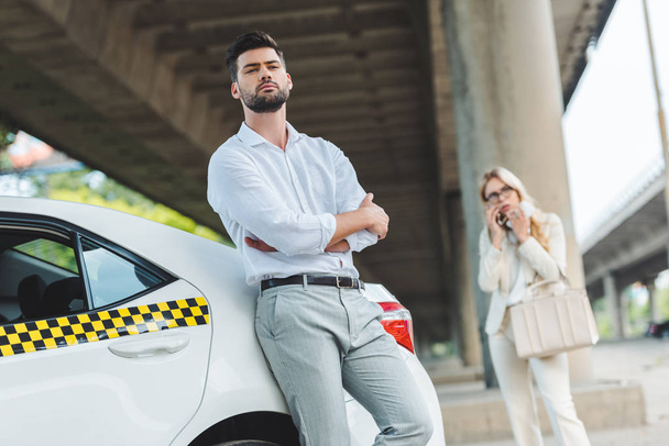 низький кут зору молодого чоловіка з схрещеними руками, що спираються на автомобіль таксі і дивиться на камеру, поки жінка стоїть позаду
 - Фото, зображення