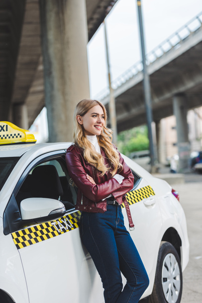 beautiful blonde girl standing with crossed arms near taxi cab and looking away - Foto, imagen