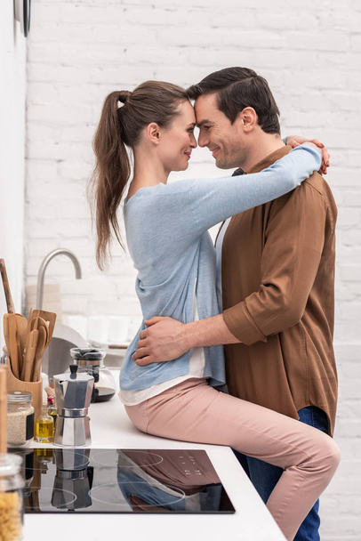 happy passionate couple embracing on kitchen while girlfriend sitting on table - Photo, Image