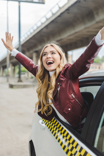 cheerful blonde girl raising hands and looking away through taxi window - 写真・画像