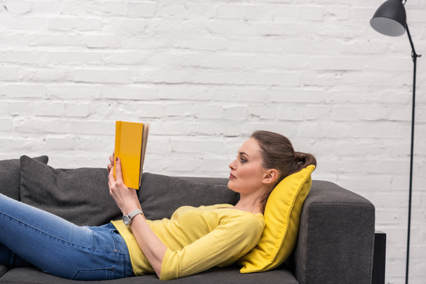 вид збоку дорослої жінки читає книгу, лежачи на дивані вдома
 - Фото, зображення