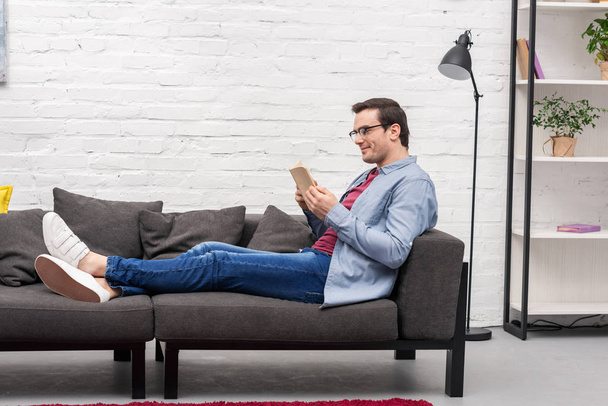 side view of happy adult man reading book on couch at home - Photo, Image