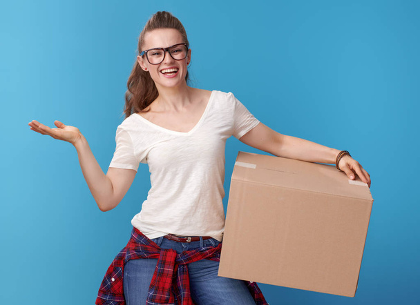 smiling active woman in white shirt with a cardboard box presenting something on empty palm isolated on blue - Zdjęcie, obraz