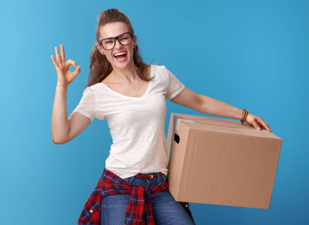 happy modern woman in white shirt with a cardboard box showing ok gesture on blue background - Foto, imagen