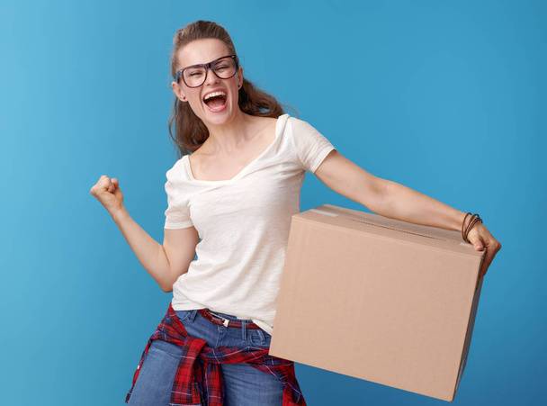 happy young woman in white shirt with a cardboard box rejoicing on blue background - Foto, imagen