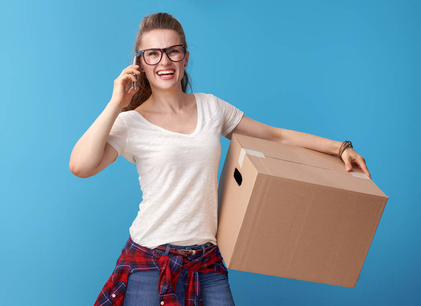 usměvavá mladá žena v bílé košili s lepenkovou krabicí mluvit na mobilním telefonu proti modrým pozadím - Fotografie, Obrázek