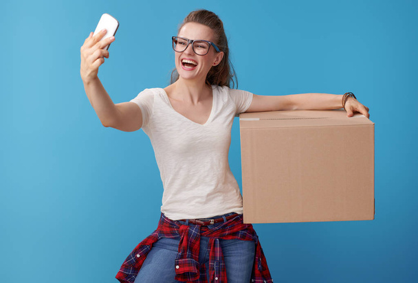 happy active woman in white shirt with a cardboard box taking selfie with cellphone against blue background - Фото, изображение