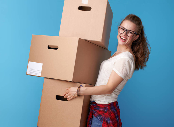 happy modern woman in white shirt holding a stack of cardboard boxes isolated on blue background - Фото, изображение
