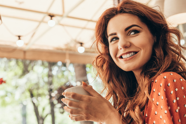 вид сбоку улыбающейся женщины с чашкой ароматного кофе в кафе
 - Фото, изображение