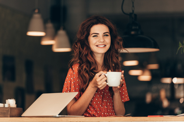 portrait of smiling woman with cup of coffee at table with laptop in cafe - Photo, Image