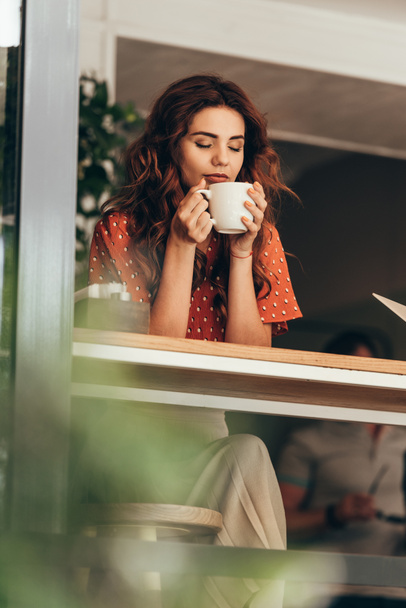 retrato de mujer hermosa con el ojo cerrado sosteniendo taza de café aromático en las manos en la cafetería
 - Foto, Imagen