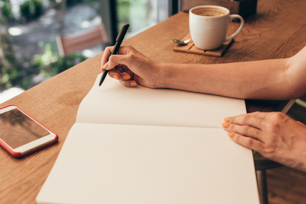 обрізаний знімок блогера, який робить нотатки в блокноті за столом з чашкою кави в кав'ярні
 - Фото, зображення