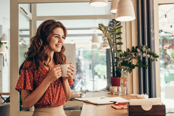 sonriente mujer joven sosteniendo taza de café en la mesa con cuaderno en la cafetería
 - Foto, Imagen