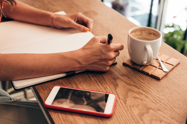 częściowy widok blogger, robiąc notatki w notesie przy stole z kubkiem kawy w kawiarni - Zdjęcie, obraz