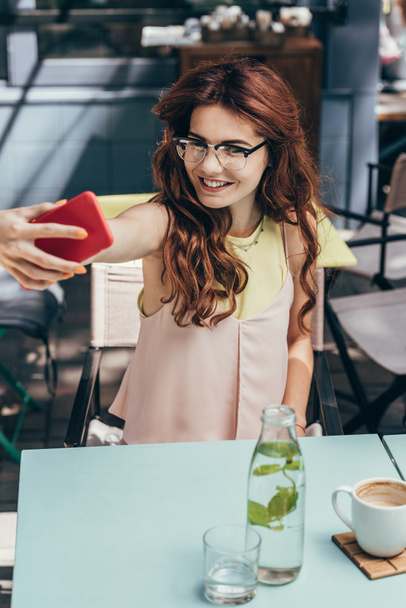 portrait of smiling woman in eyeglasses taking selfie on smartphone in cafe - Фото, изображение