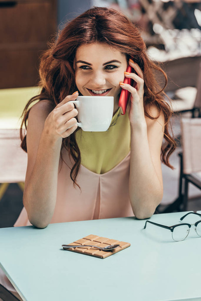 portrait of smiling woman with cup of coffee talking on smartphone in cafe - Foto, Imagem