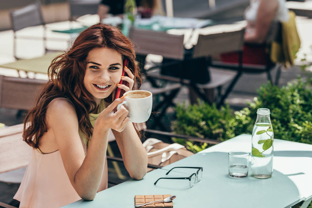 portrait of smiling woman with cup of coffee talking on smartphone in cafe - Photo, Image