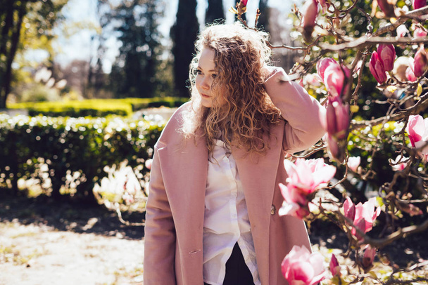 lockige elegante Frau im trendigen rosafarbenen Mantel, die durch den Park spaziert und die Wärme und den Frühling genießt - Foto, Bild