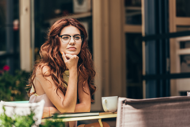 Porträt einer nachdenklichen Geschäftsfrau mit Brille am Tisch mit Tasse Kaffee und Notizbuch im Café - Foto, Bild