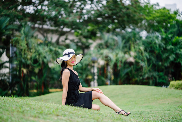 Portrait of Asian Chinese woman in an elegant  black dress posing with a white broad-brimmed hat during the day in a park in Asia. - Φωτογραφία, εικόνα