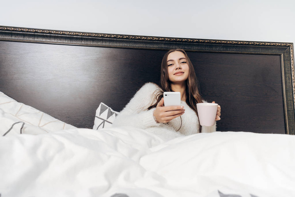 beautiful young girl in pajamas lying in bed in the evening, resting after a hard day's work and holding a smartphone - 写真・画像