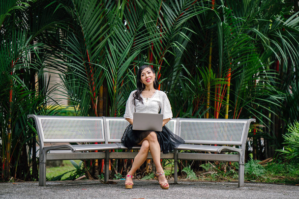 An Asian woman is sitting on a metal bench with trees in the background and is working on her laptop computer. She is middle aged, thin and is dressed in office attire. - Fotoğraf, Görsel