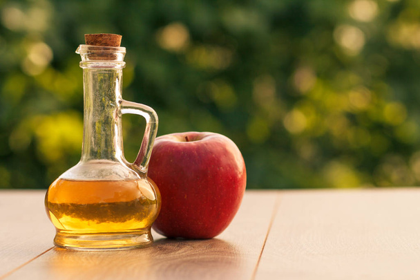 Яблучний оцет у скляній пляшці з пробкою та свіжим червоним яблуком на дерев'яних дошках з розмитим зеленим природним фоном. Органічна їжа для здоров'я
 - Фото, зображення