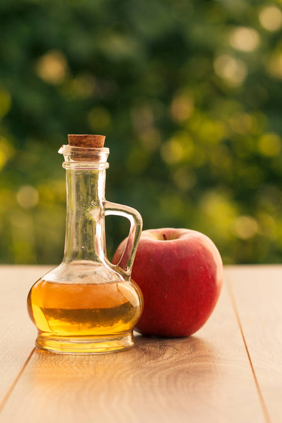 Яблочный уксус в стеклянной бутылке с пробкой и свежим красным яблоком на деревянных досках с зеленым естественным фоном. Органическое питание для здоровья
 - Фото, изображение