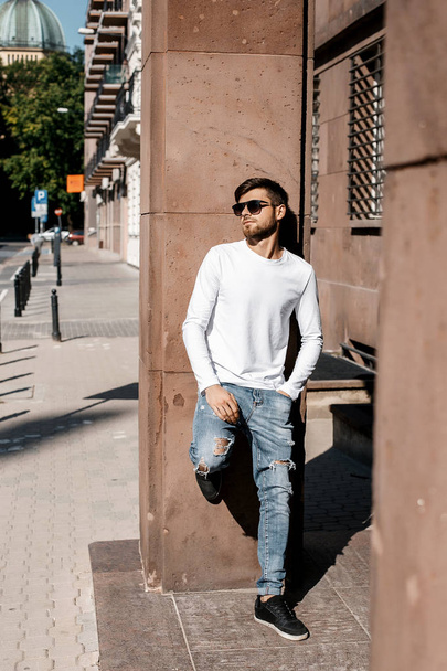 Un jeune homme parcourt les rues de la ville. Garçon en lunettes de soleil Homme élégant près de l'immeuble. Il est assis sur un fouet. Barbe et les cheveux dans un gars élégant
. - Photo, image