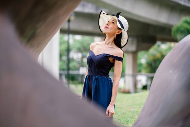 Portrait of Asian Chinese woman in an elegant  blue dress posing with a white broad-brimmed hat during the day in a park in Asia. - 写真・画像