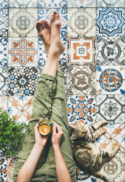 Womans legs in cosy linen pants, plant, cat and cup of coffee in hand over colorful moroccan tile floor - Foto, Bild
