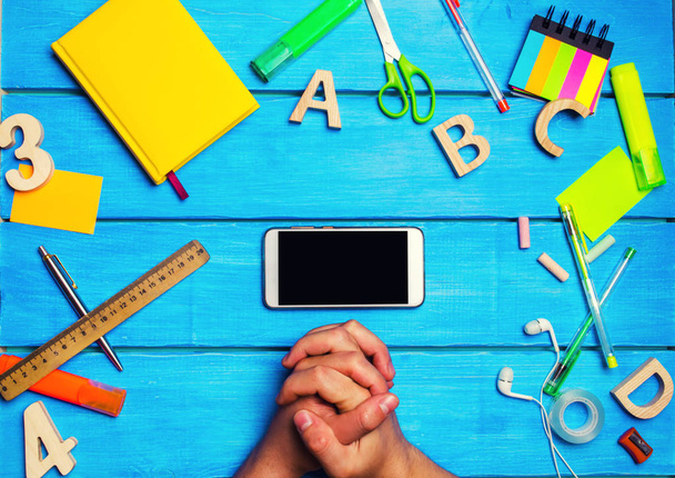 The student crossed his hands in front of a mobile phone in the workplace. A place for text. A student is looking at a mobile phone. Creative clutter on a blue wooden background. concept of education. - Zdjęcie, obraz