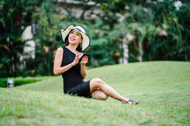 Porträt einer asiatischen Chinesin in einem eleganten schwarzen Kleid, die tagsüber in einem Park in Asien mit einem weißen breitkrempigen Hut posiert. - Foto, Bild