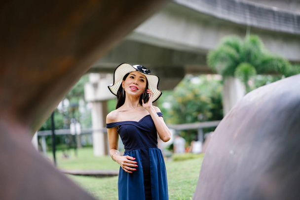 Portrait of a stylish Asian woman in a blue dress and white hat in  park with smartphone. - Фото, зображення