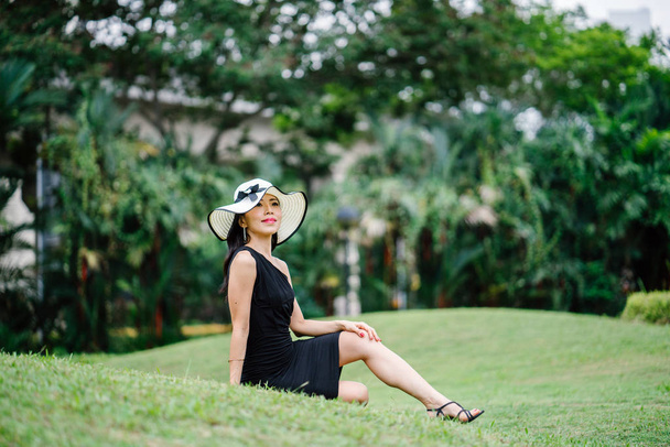 Portrait of Asian Chinese woman in an elegant  black dress posing with a white broad-brimmed hat during the day in a park in Asia. - Valokuva, kuva