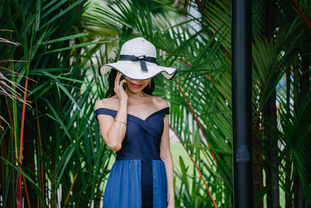Portrait of a stylish Asian woman in a blue dress and white hat in  park with smartphone. - Fotografie, Obrázek