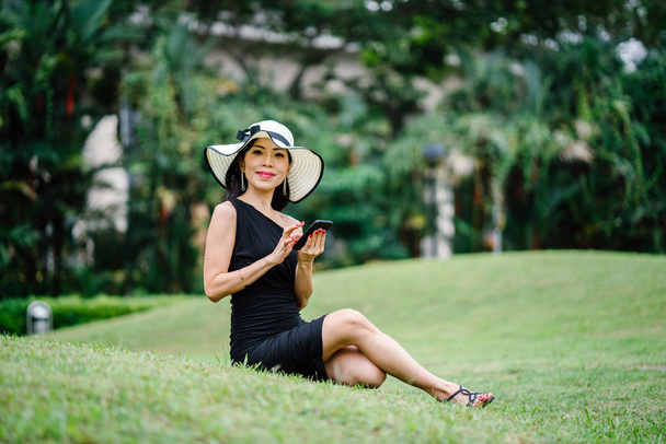 Portrait of a stylish Asian woman in black dress and white hat in  park with smartphone. - Фото, зображення