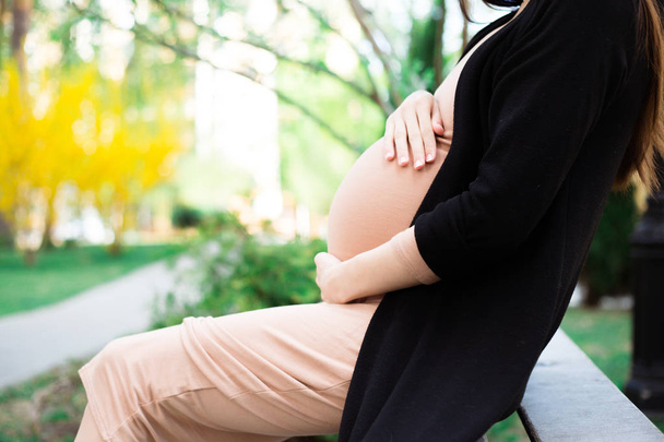 Partial view of pregnant woman in beige dress holding belly sitting on bench in park - Foto, imagen
