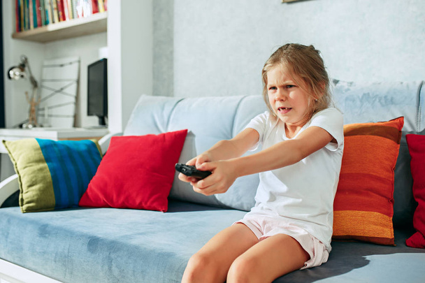 Little casual girl watching tv at home. Female kid sitting on sofa with TV remote and switching channels - Fotoğraf, Görsel